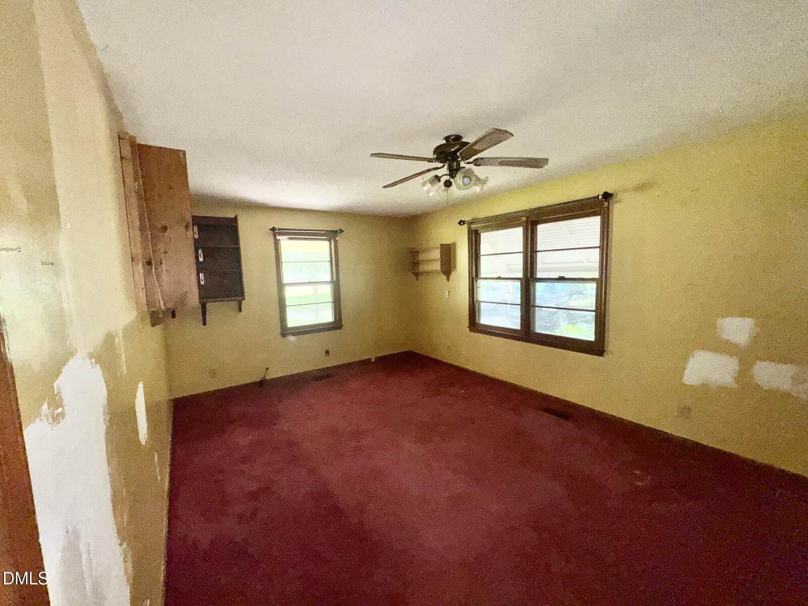 Room with yellow walls, red carpet, ceiling fan with lights, two windows with wooden blinds, wooden shelves on walls, and visible patches on the right wall. Room appears empty and unfurnished.