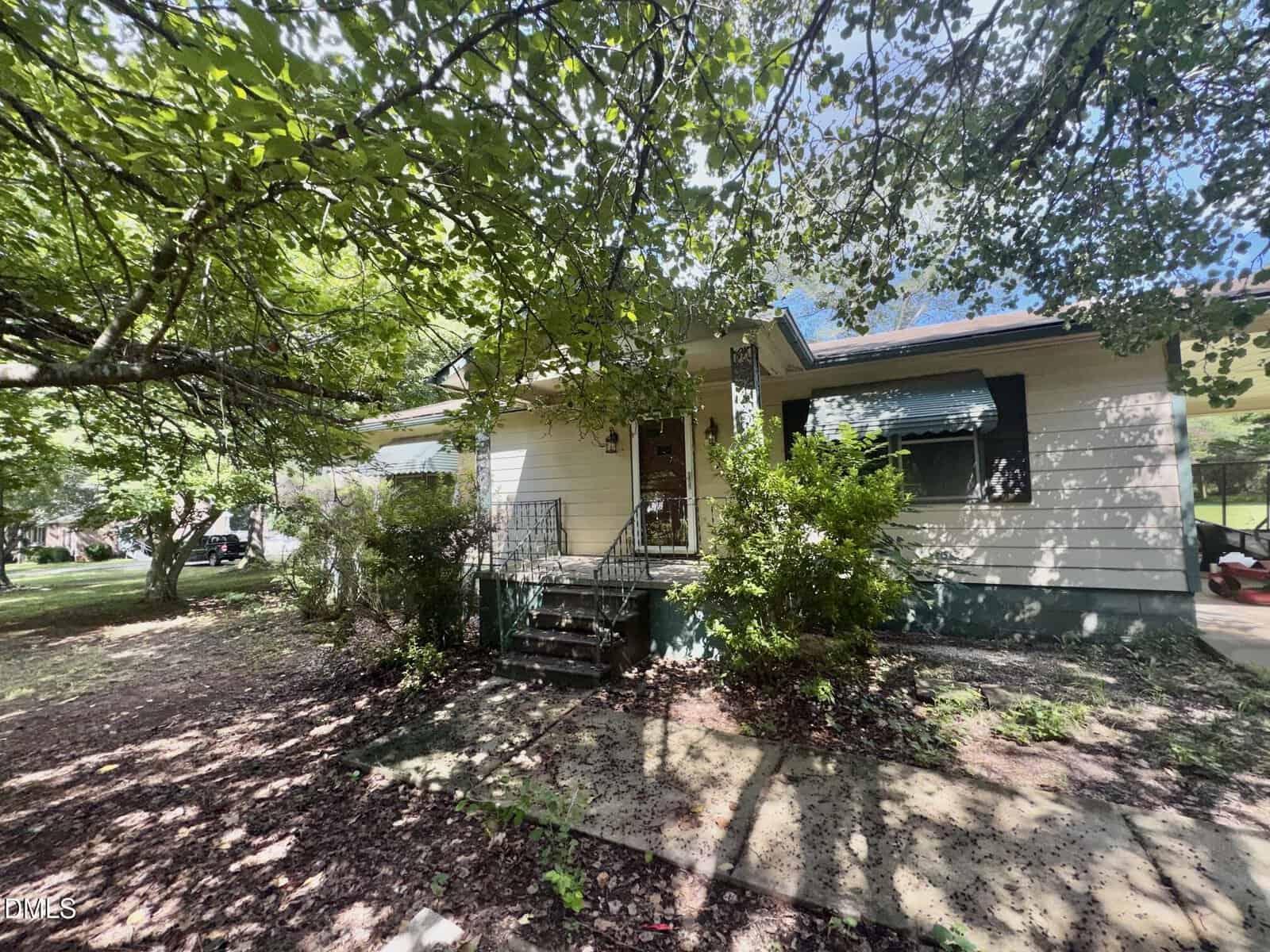 A single-story house with light-colored siding, green trim, a front porch with steps, and shaded by large trees stands invitingly at 362 Country Acres Road in Henderson. Bushes and grass surround the entryway, while sunlight filters through the branches onto the house.