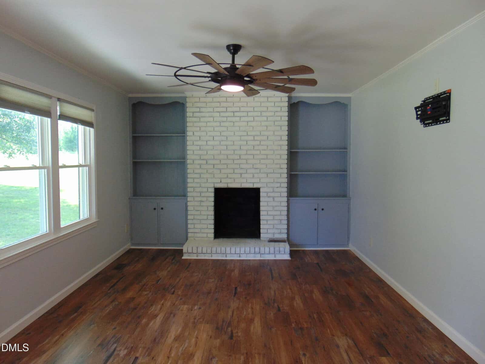 A living room with wood flooring, white brick fireplace, built-in shelves and cabinets painted gray, large ceiling fan, windows on the left, and a small wall-mounted item on the right wall.