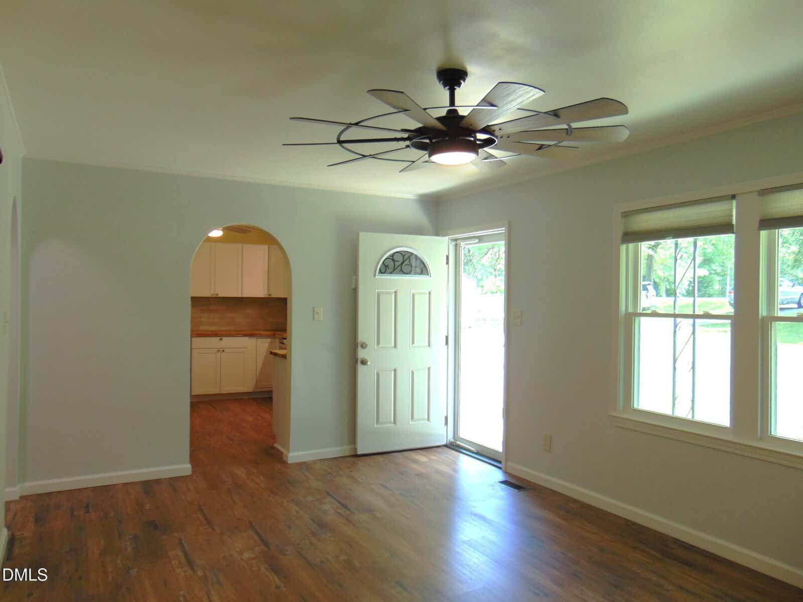 A bright living room with wood flooring, a large ceiling fan, an open front door, and double windows. An arched doorway leads to a kitchen with white cabinets and a tile backsplash.