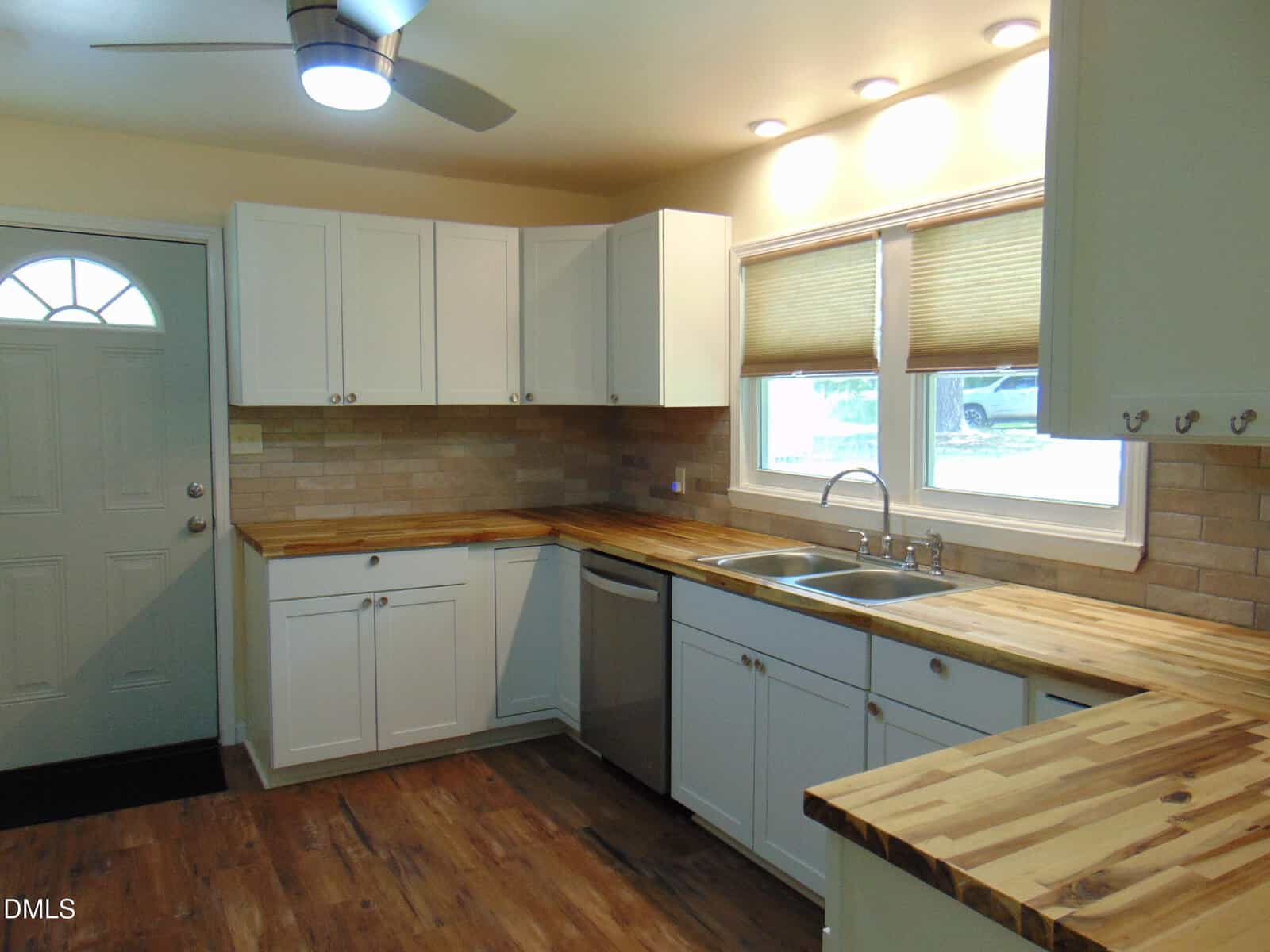 Modern kitchen with white cabinets, wood countertops, beige tile backsplash, stainless steel sink and dishwasher, under-cabinet lighting, a window above the sink, a ceiling fan, and a door to the left.