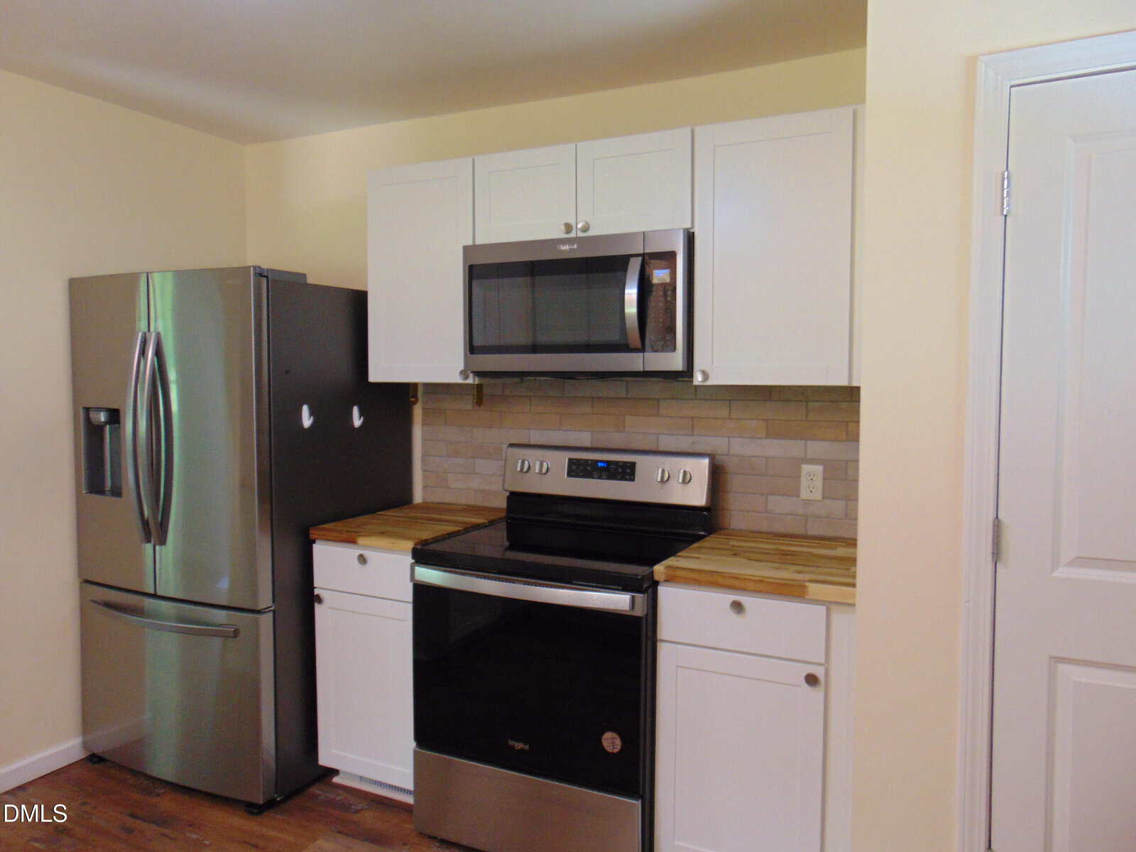 A kitchen with a stainless steel refrigerator, electric stove, and microwave. White cabinets and drawers are above and beside the appliances. The countertop has a wood finish and the backsplash is made of light brown tiles.