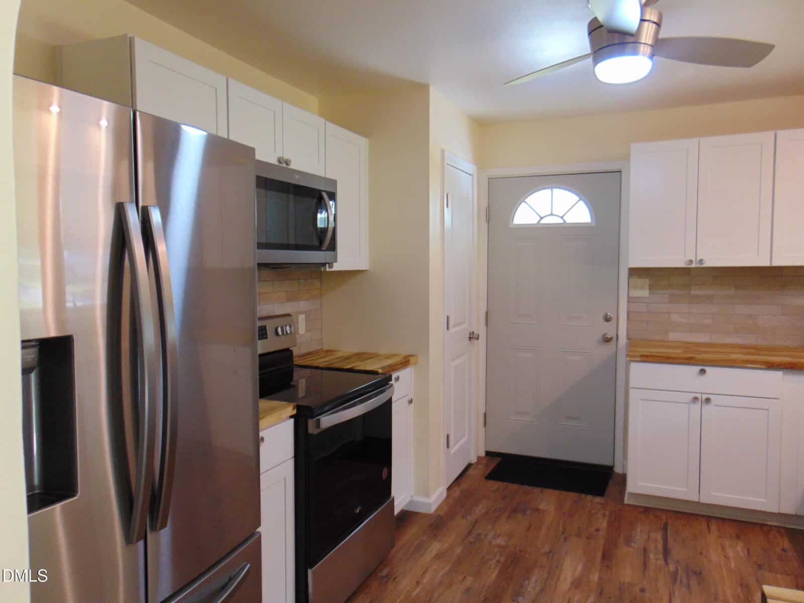 Modern kitchen with stainless steel appliances, white cabinets, wood countertops, tile backsplash, and a gray door with a round window. Ceiling fan with light and wood-style flooring are also visible.