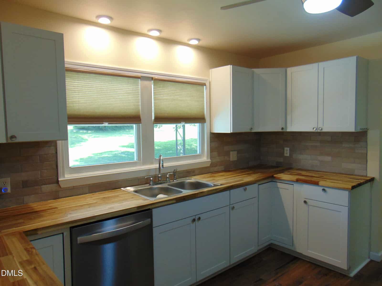 Modern kitchen with white cabinets, wood countertops, stainless steel dishwasher, double sink, tan tile backsplash, and two windows with beige shades. Recessed lighting is installed above the sink area.