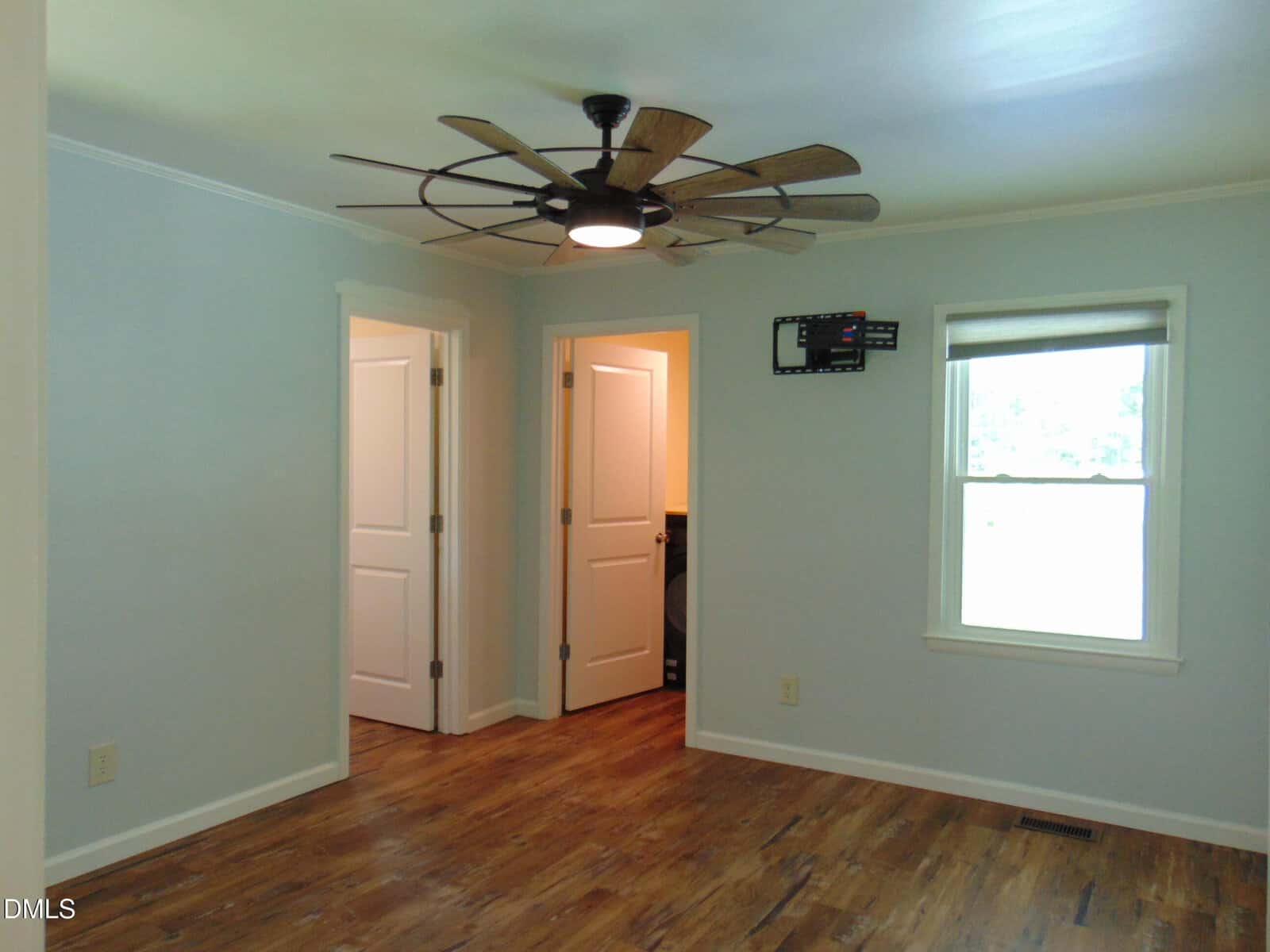 A bright, empty room with light blue walls, wooden floor, large ceiling fan, two white doors, a window with a blind, and a wall-mounted TV bracket above an outlet.