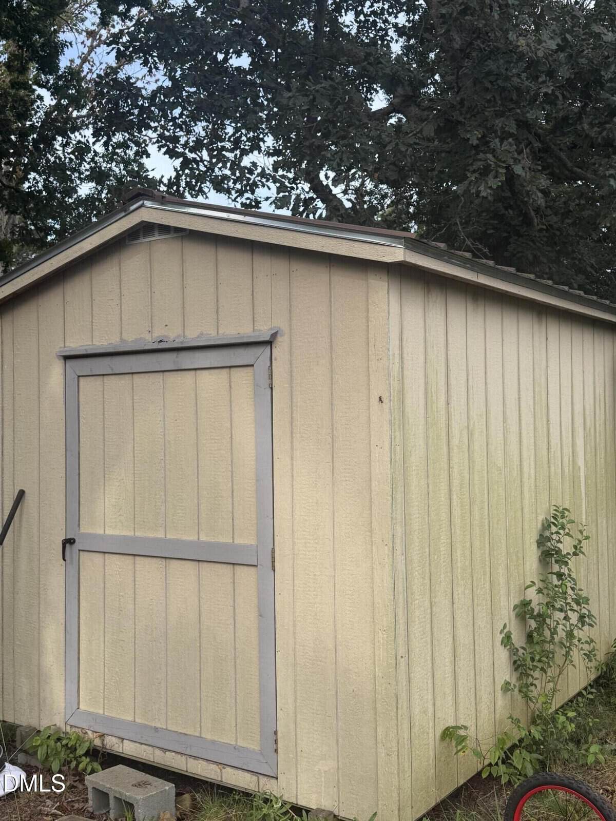 A beige wooden shed with a single door and a slanted metal roof is shown. Some greenery is growing near the shed, and trees with dense foliage are visible in the background. The shed appears weathered.
