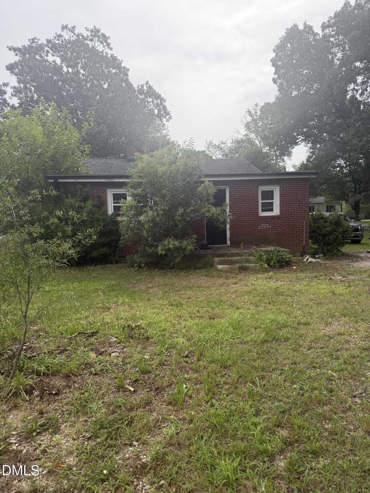 A small, single-story brick house with two front windows is partially obscured by overgrown bushes. The yard is grassy and unkempt, with trees and shrubs surrounding the house under a cloudy sky.