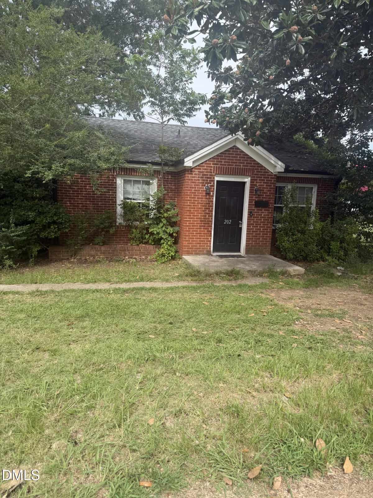 A small, one-story red brick house sits at 202 Mitchell Avenue in Franklinton, featuring a black front door, two windows, and a gabled roof. The yard is grassy with some patches of dirt, while bushes and trees surround the house.