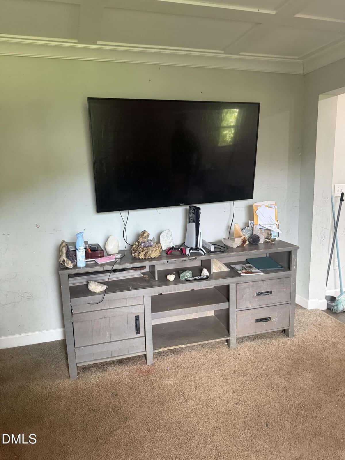 A flat-screen TV is mounted on a light gray wall above a wooden entertainment console, which holds rocks, papers, cleaning spray, and miscellaneous items. The carpet is beige, and the room appears cluttered.
