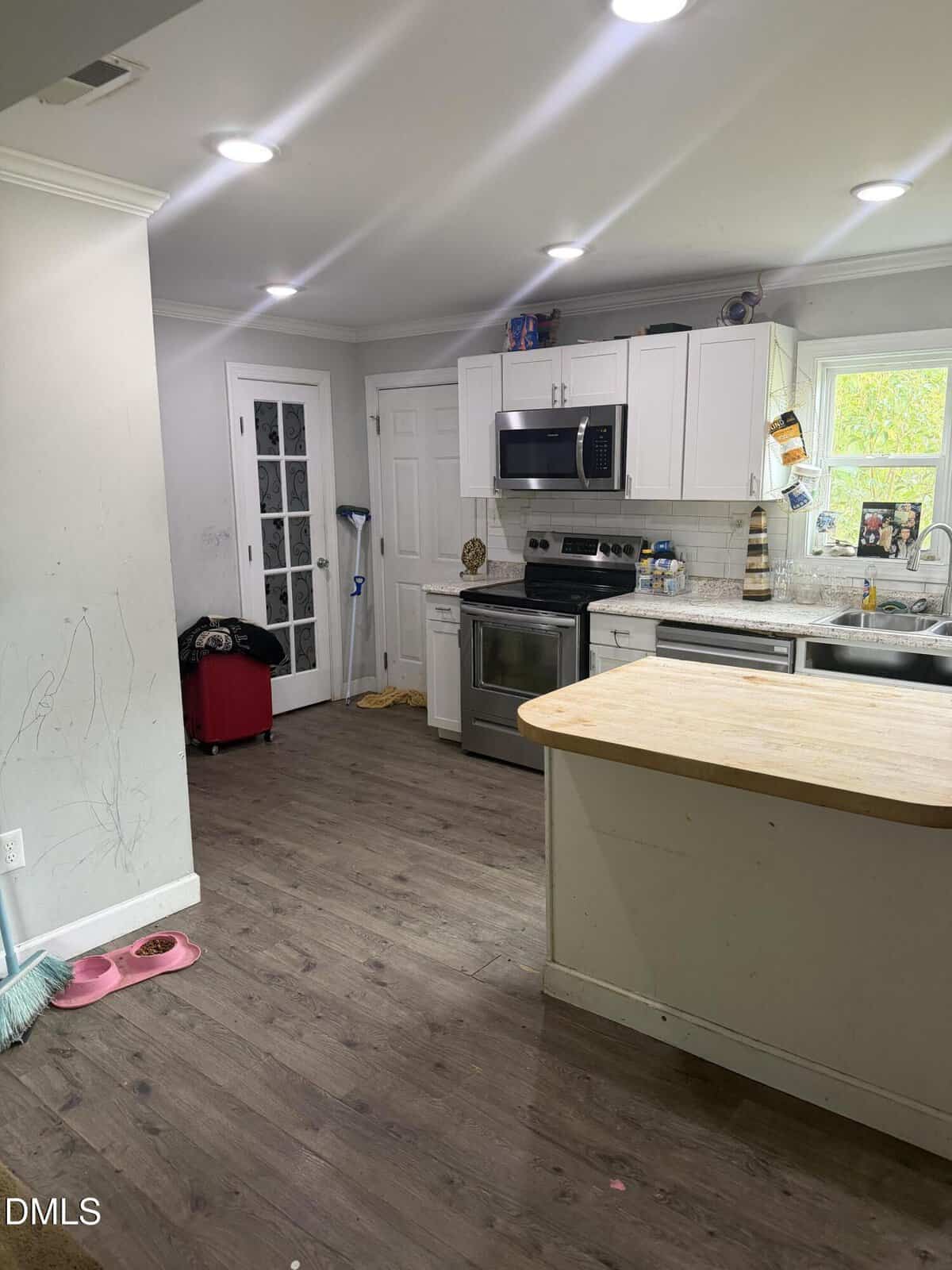 A kitchen with white cabinets, stainless steel appliances, wood floors, and a small island. A mop, red suitcase, and cat drawing are visible, along with clutter on counters and marks on the wall.