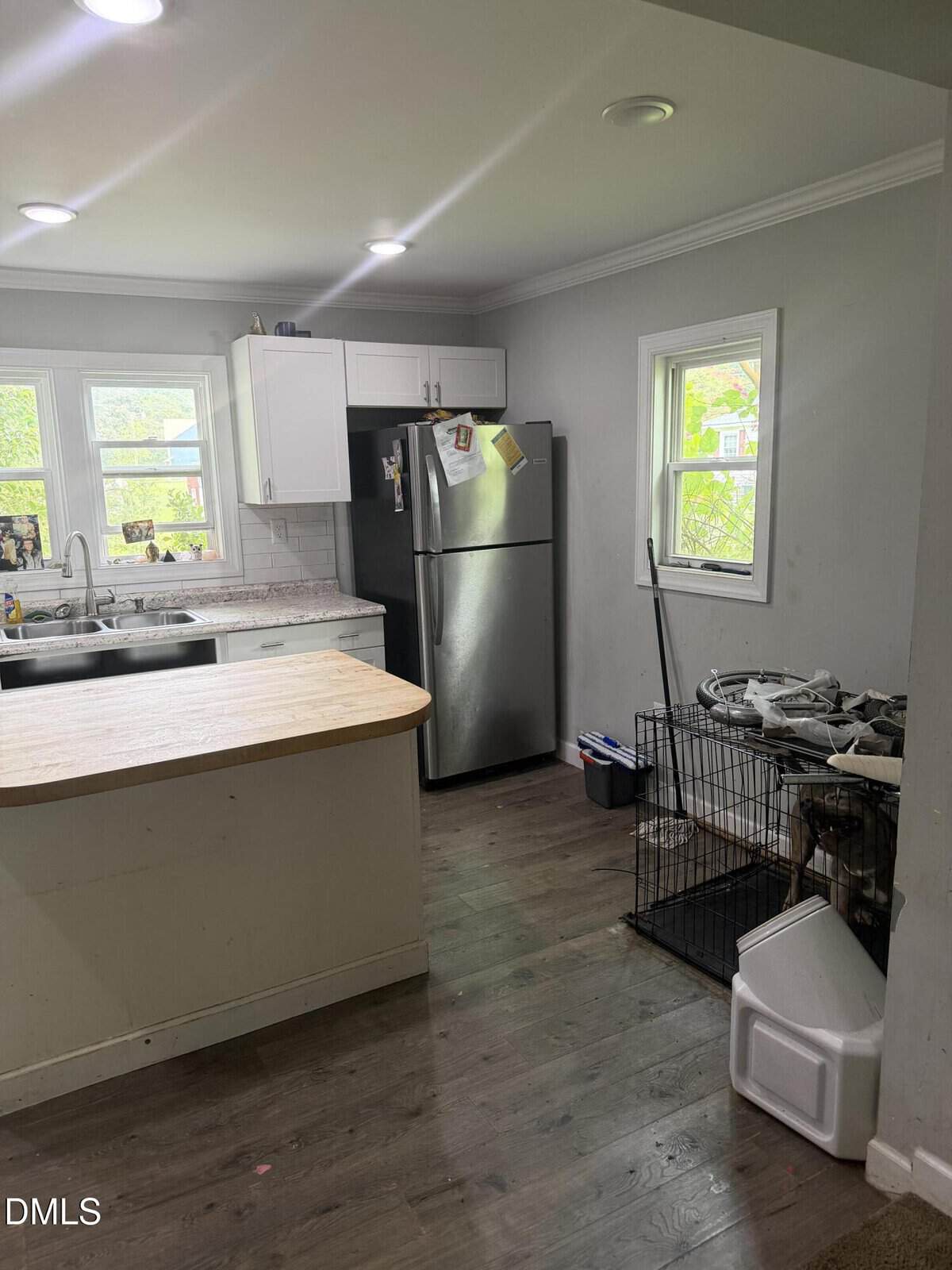 A kitchen with white cabinets, a stainless steel fridge, a wooden island, and a window. A dog crate with a dog inside sits by the fridge. Various kitchen items and a trash bin are visible on the right.