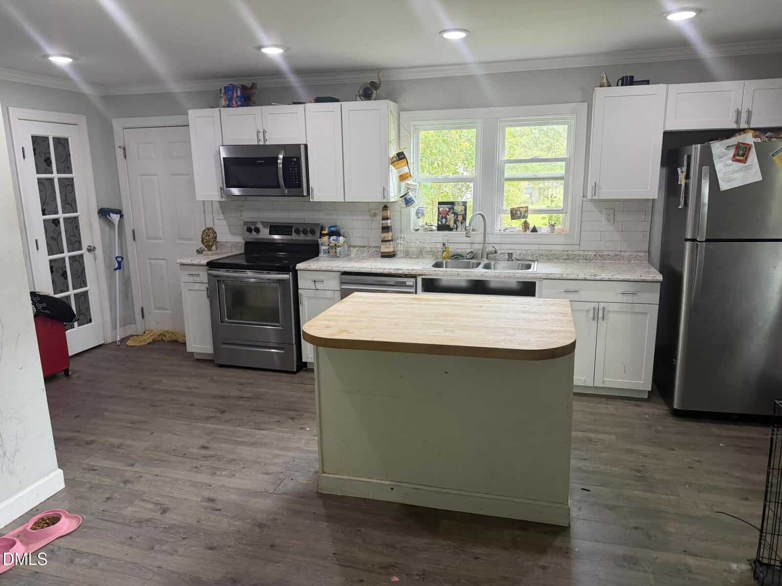 A modern kitchen with white cabinets, stainless steel appliances, a small center island, and two windows above the sink. Items and magnets are on the fridge, and cleaning supplies are visible near the door.
