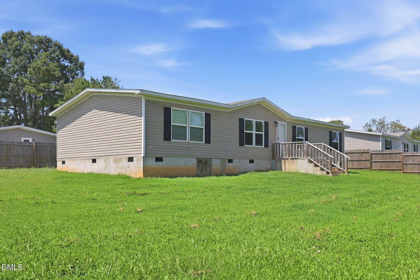 A single-story manufactured home with beige siding, white trim, and black shutters sits on a grassy lot. The house features a small wooden porch with steps and a fenced backyard. The sky is bright and mostly clear.