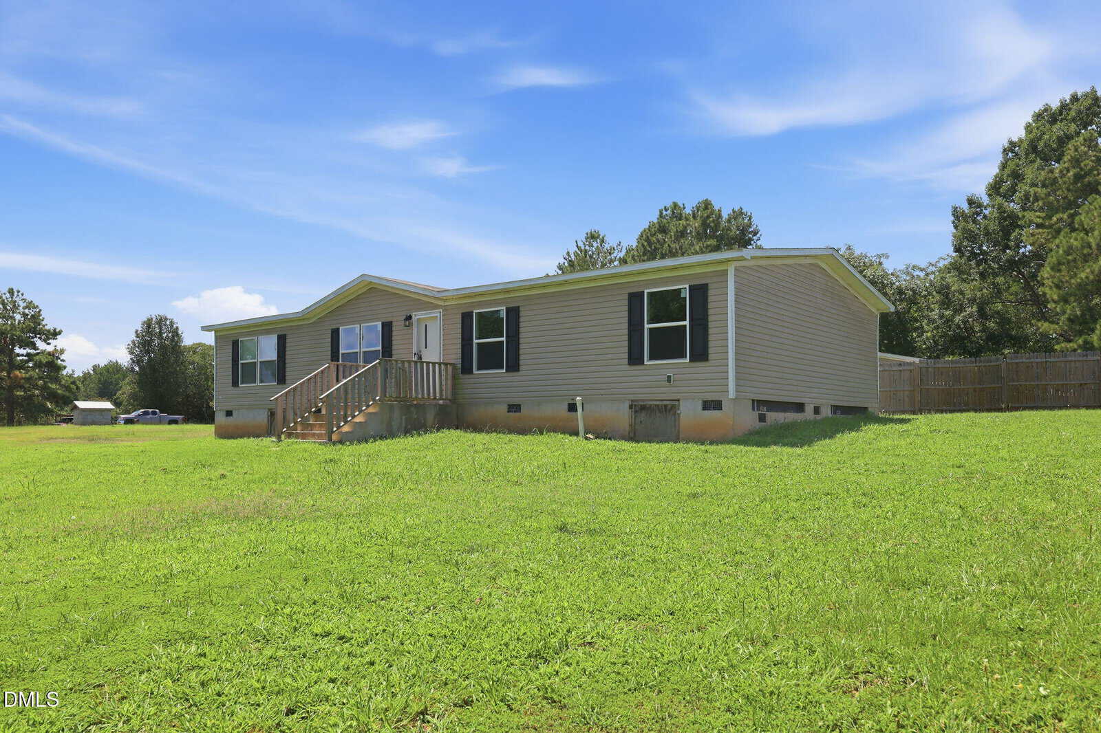 Single-story manufactured home with beige siding, white trim, a small front porch with stairs, and a large grassy yard. Trees and a wooden fence are visible in the background under a blue sky with wispy clouds.