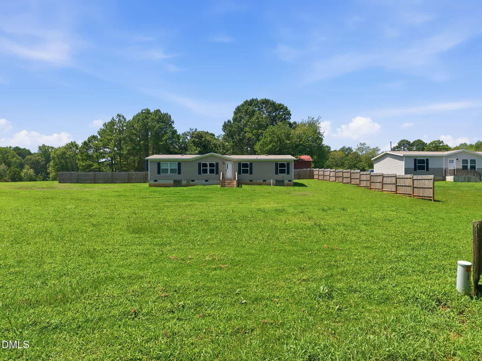 A single-story manufactured home sits in the center of a large, grassy yard with a wooden fence and trees in the background under a blue sky with light clouds. Another similar home is visible to the right.