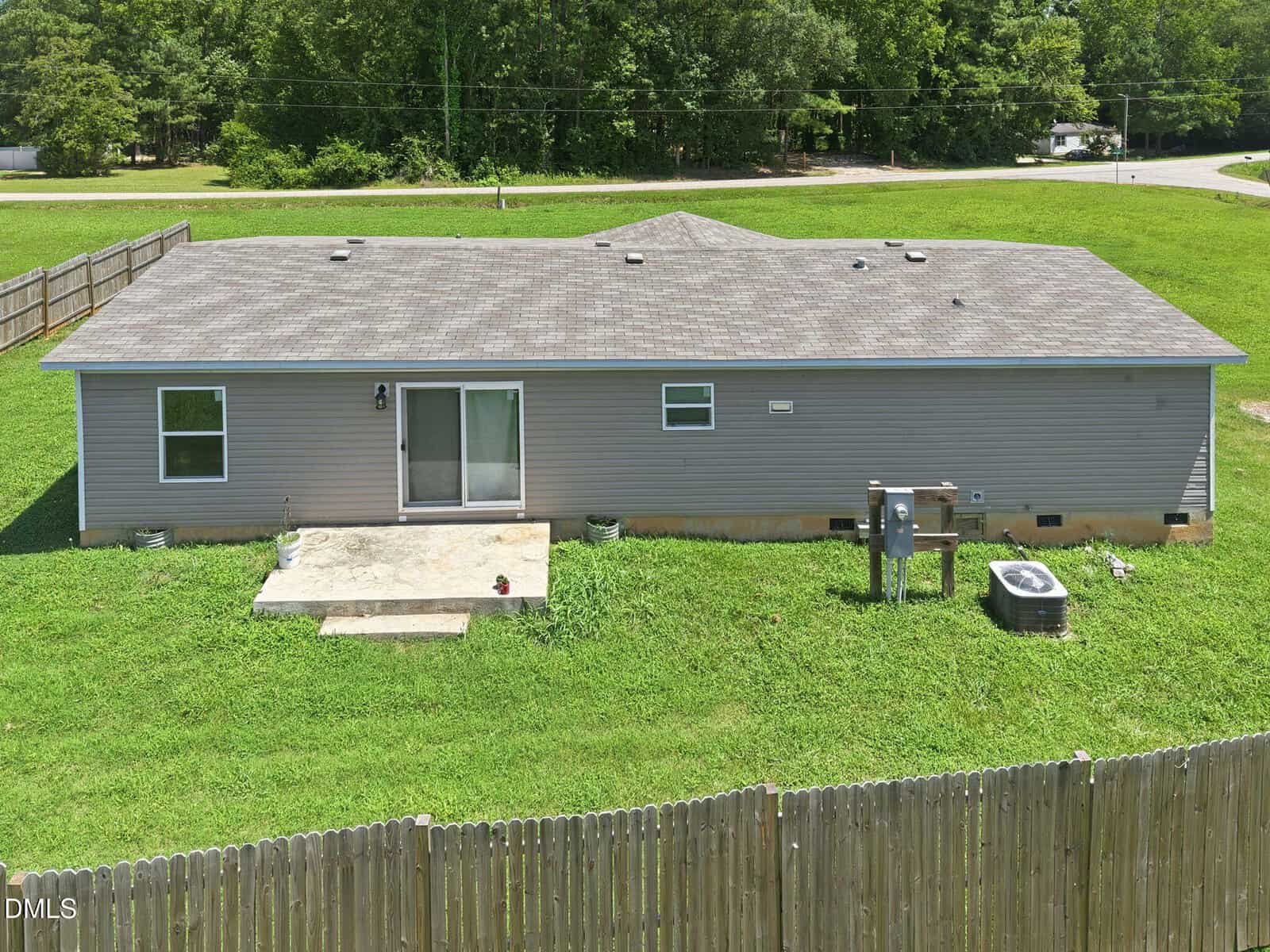 Single-story house with gray siding, three visible windows, and a small concrete patio in back. The yard is grassy and fenced, with an air conditioning unit and utility meters next to the house. Trees are visible in the background.