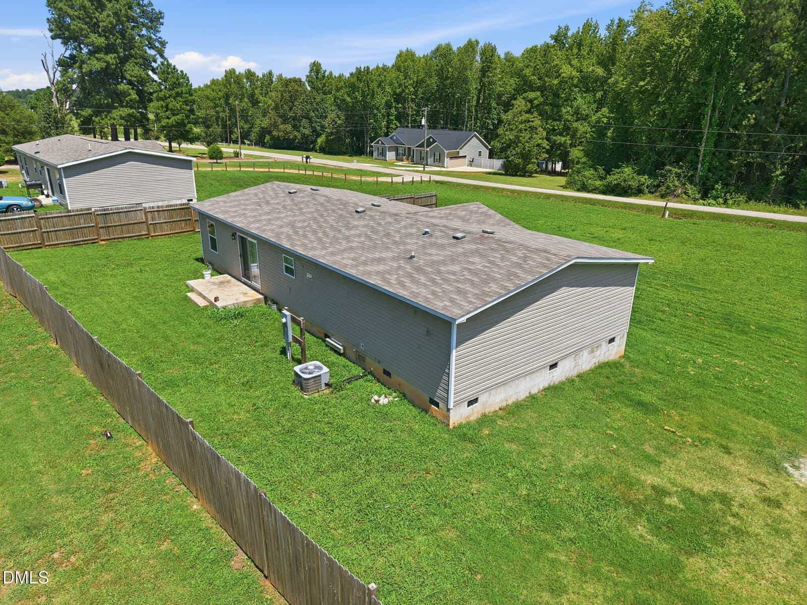 A light gray single-story house with a fenced backyard, set on a large grassy lot, surrounded by trees and neighboring houses under a blue sky.