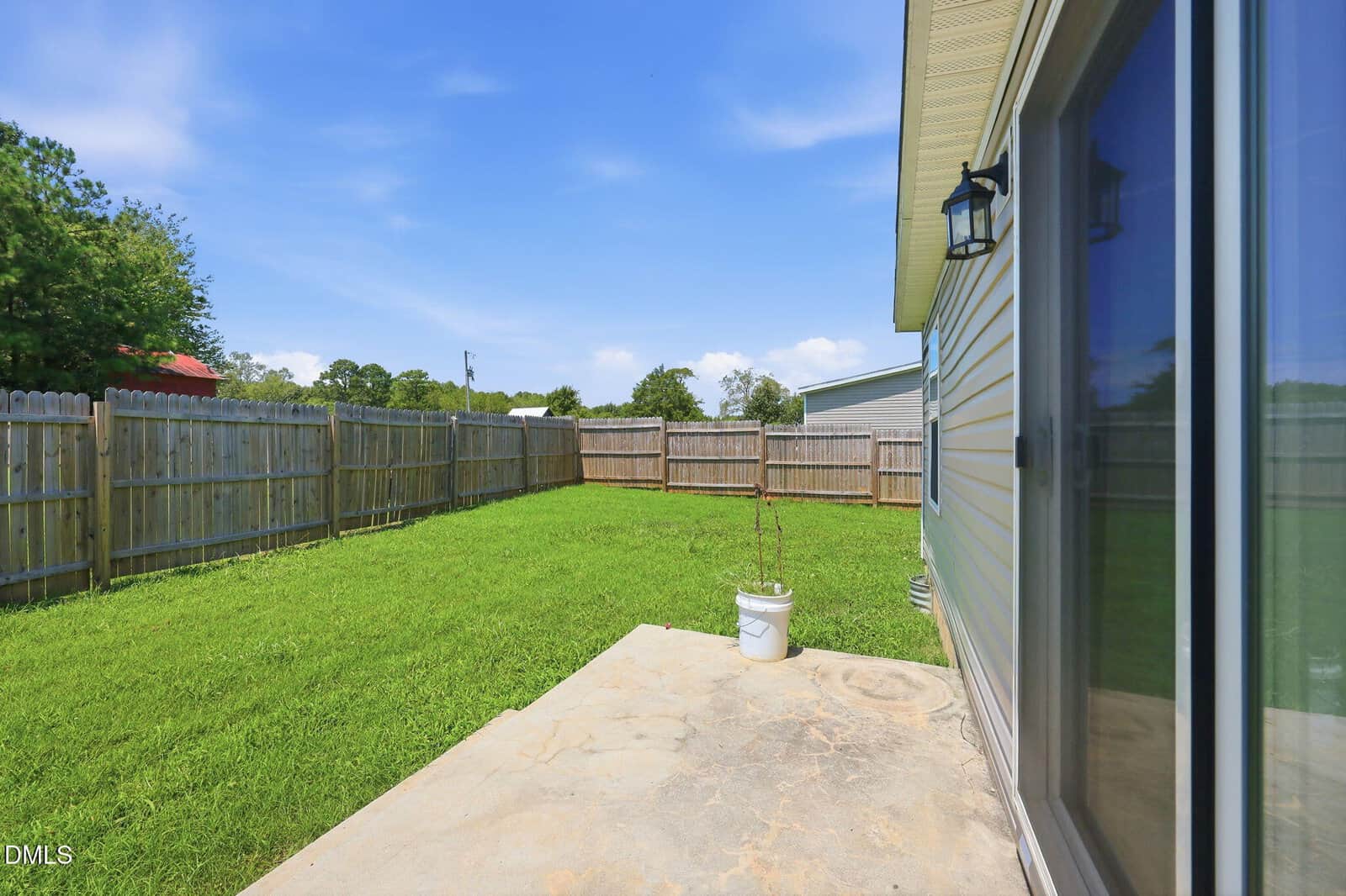 A backyard with a wooden fence, green grass, a small potted plant on a concrete patio, and a house with sliding glass doors and an outdoor wall light. Blue sky with a few clouds overhead.