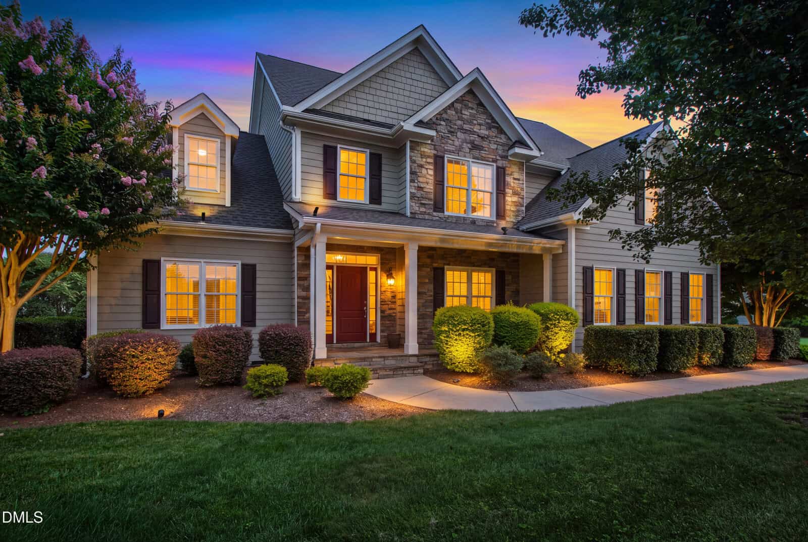 Two-story suburban house with gray siding, stone accents, front porch, and illuminated windows at dusk. The yard features neatly trimmed bushes, green lawn, and trees with evening sky in the background.