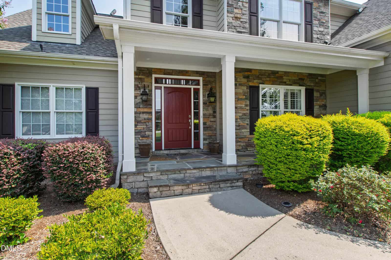 Front entrance of a house with stone and siding exterior, a covered porch with columns, a red front door, and trimmed bushes and shrubs along a curved concrete walkway.