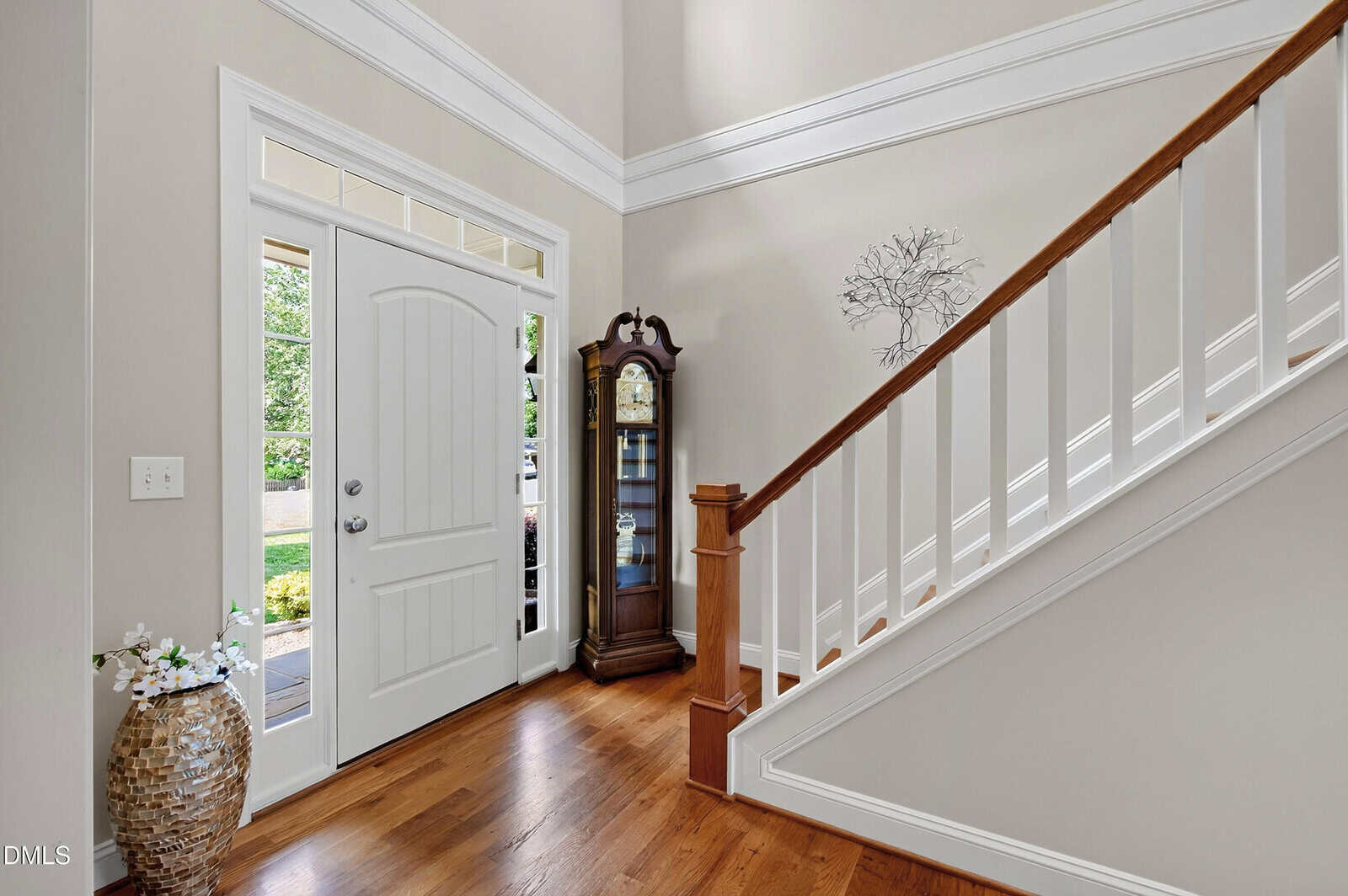 A foyer with a white front door, glass side panels, wooden floor, a grandfather clock, a large vase with twigs, and a wooden staircase with white railings. Neutral walls and natural light are present.