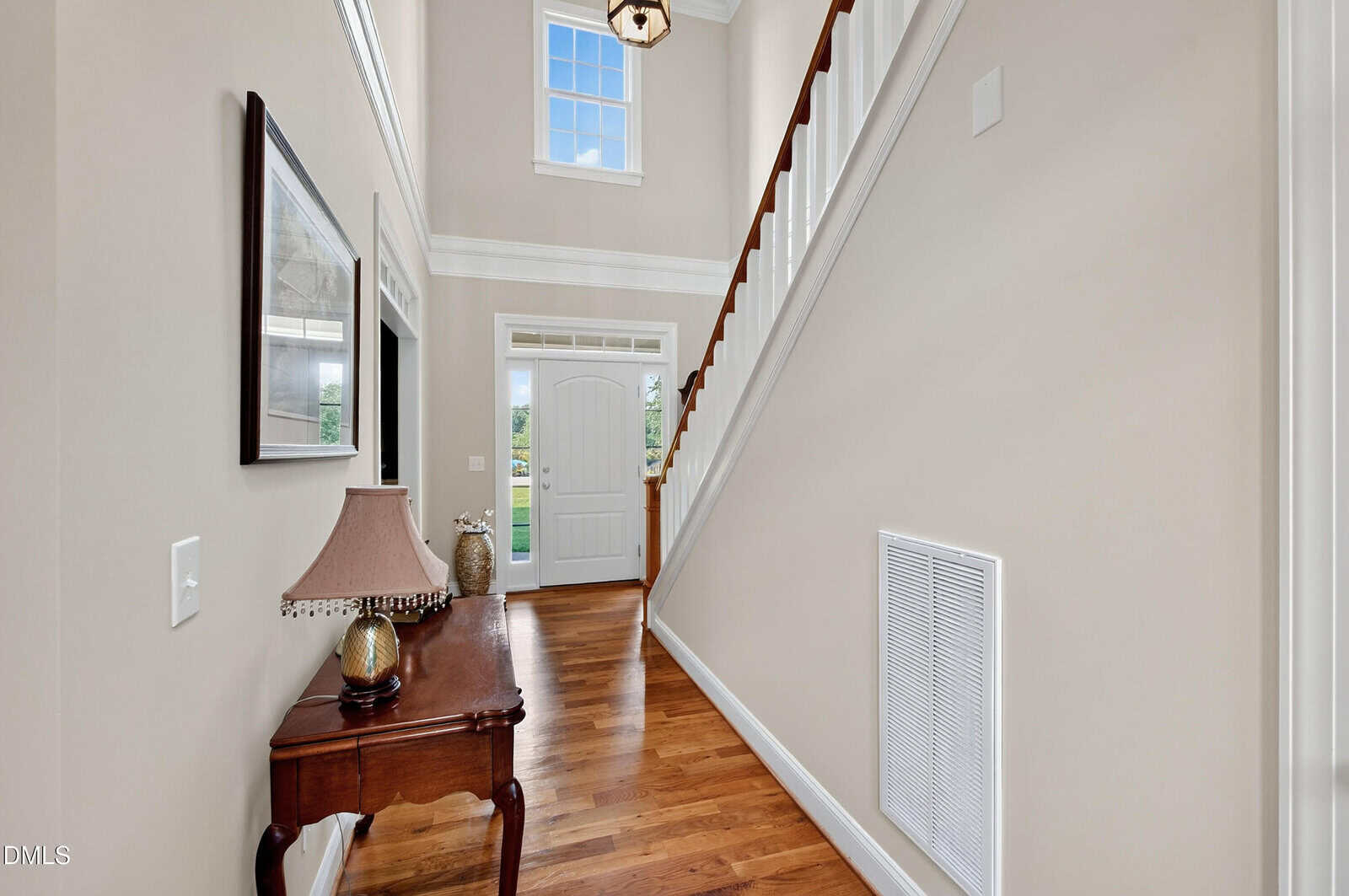 A bright hallway with wooden floors, a staircase on the right, a console table with a lamp and decor on the left, and a glass-paneled front door letting in natural light.