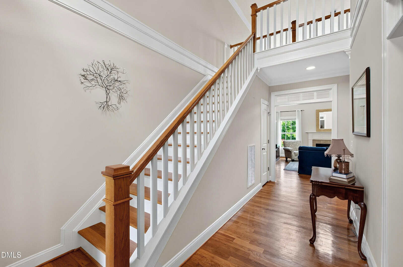 A hallway with a wooden staircase and white railings, light-colored walls, a wall art piece, a small table with a lamp, and a view into a living room with a couch and large windows.