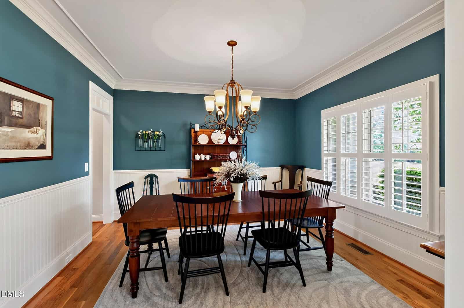 A dining room with blue walls, a wooden table, six black chairs, a chandelier, and a hutch with decor. Sunlight enters through white shuttered windows, and a rug lies beneath the table.