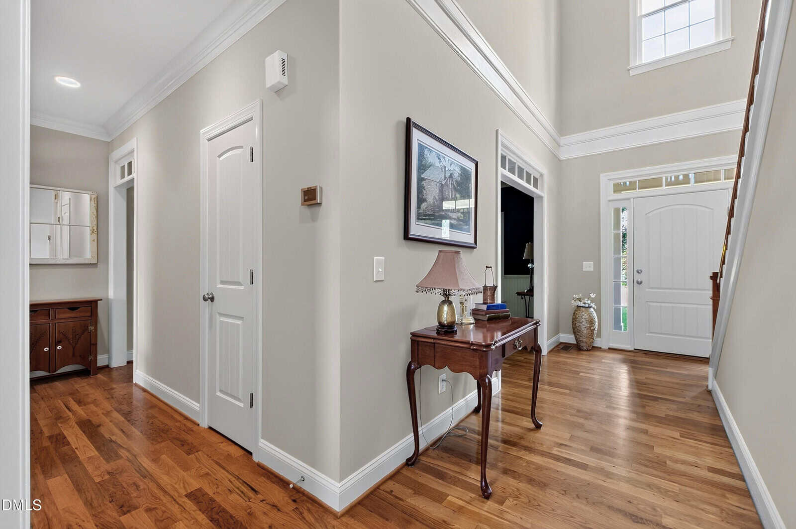 Bright entryway with light walls, hardwood floors, a white front door with glass panels, a wooden table with a lamp and books, and a staircase leading up. Natural light streams in from windows above the door.
