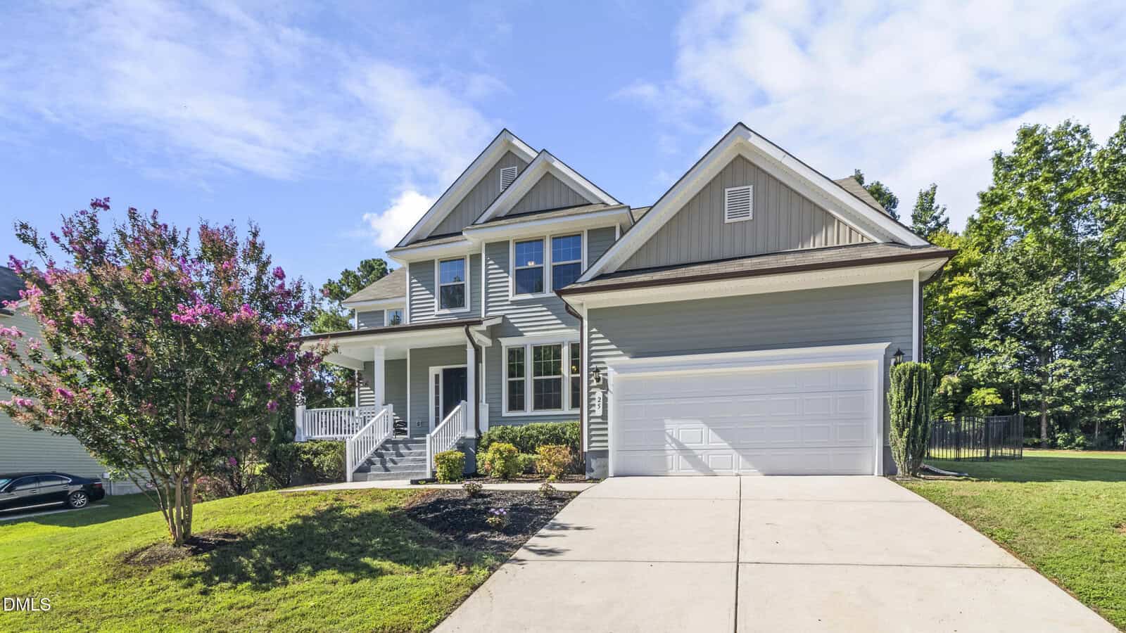 A two-story light blue house with white trim, a front porch, and a double garage. The driveway is concrete, and there is a flowering tree and green lawn in the front yard. The sky is partly cloudy.