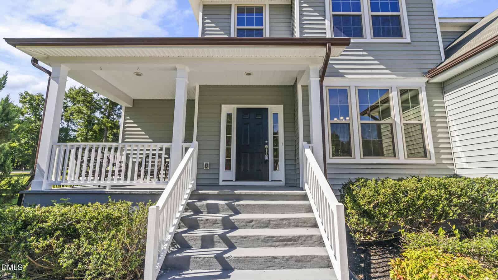 Front view of a modern house with gray siding, black front door, white trim, a small porch with white railing, steps leading up, and green bushes along the walkway.