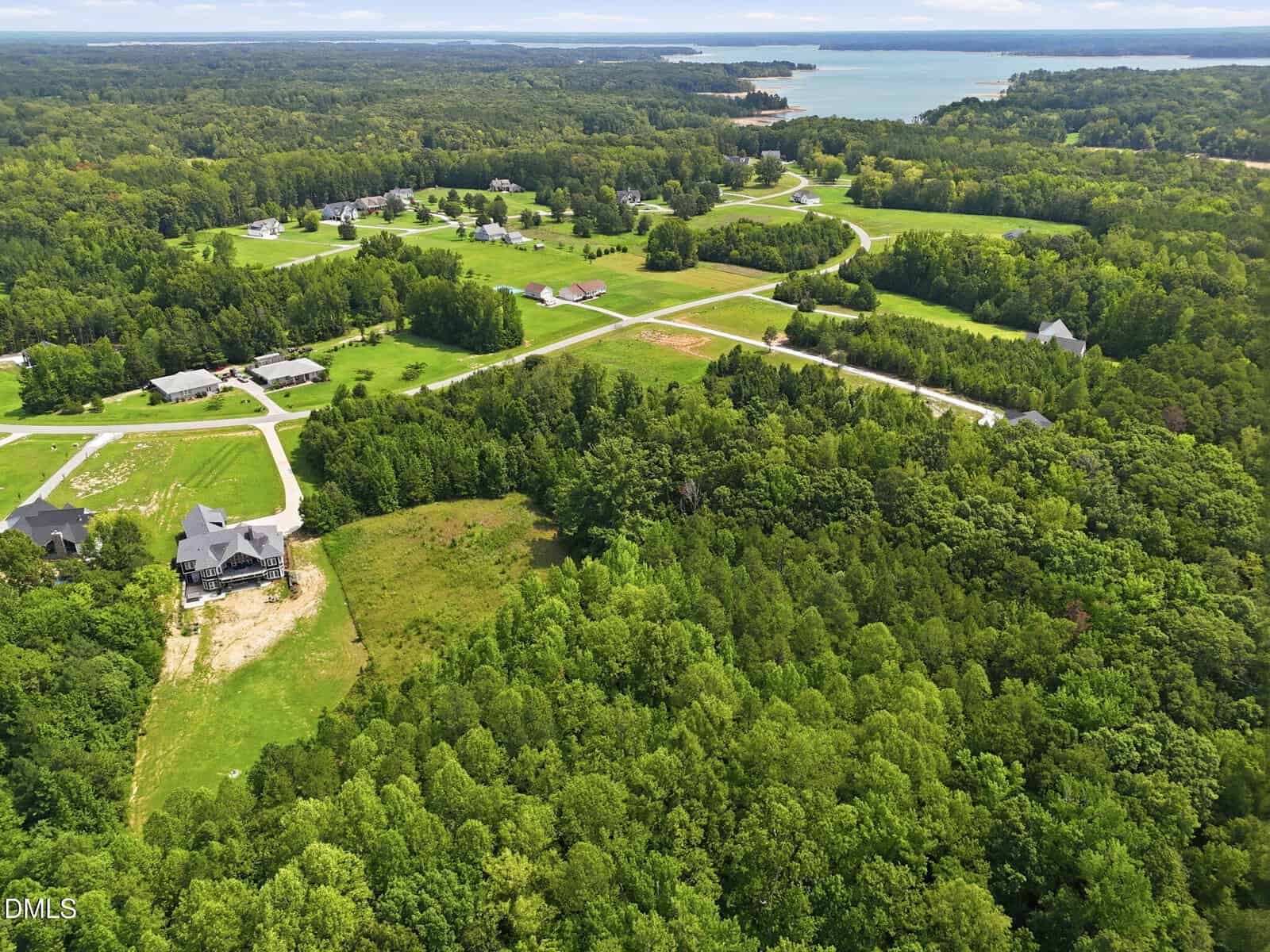 Aerial view of a rural neighborhood with scattered houses, winding roads, large green lawns, and dense forest areas. A lake is visible in the distance under a cloudy sky.