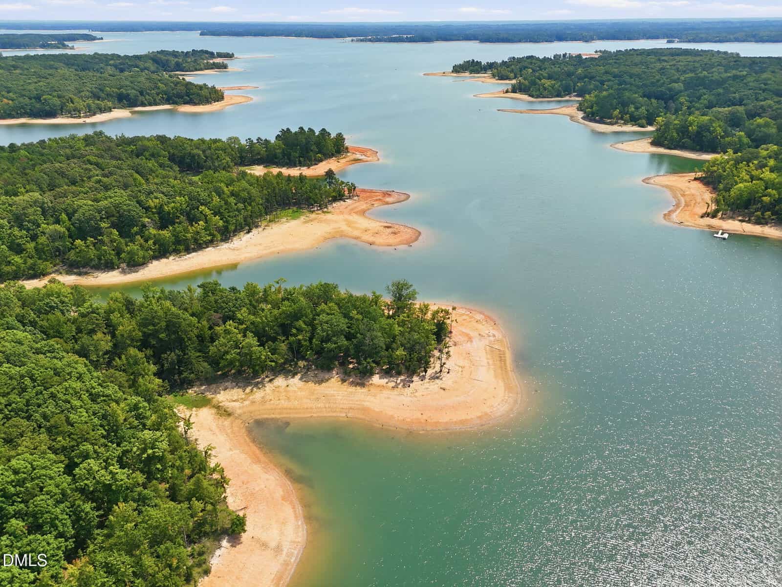 Aerial view of a lake with multiple green, tree-covered peninsulas and sandy shorelines, surrounded by calm blue water under a cloudy sky.