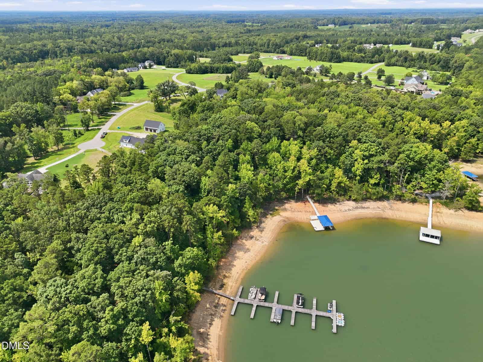 Aerial view of a green, wooded shoreline with a small marina, several docks, and a sandy beach area next to calm water. Houses and winding roads are visible further inland among open grassy areas and trees.