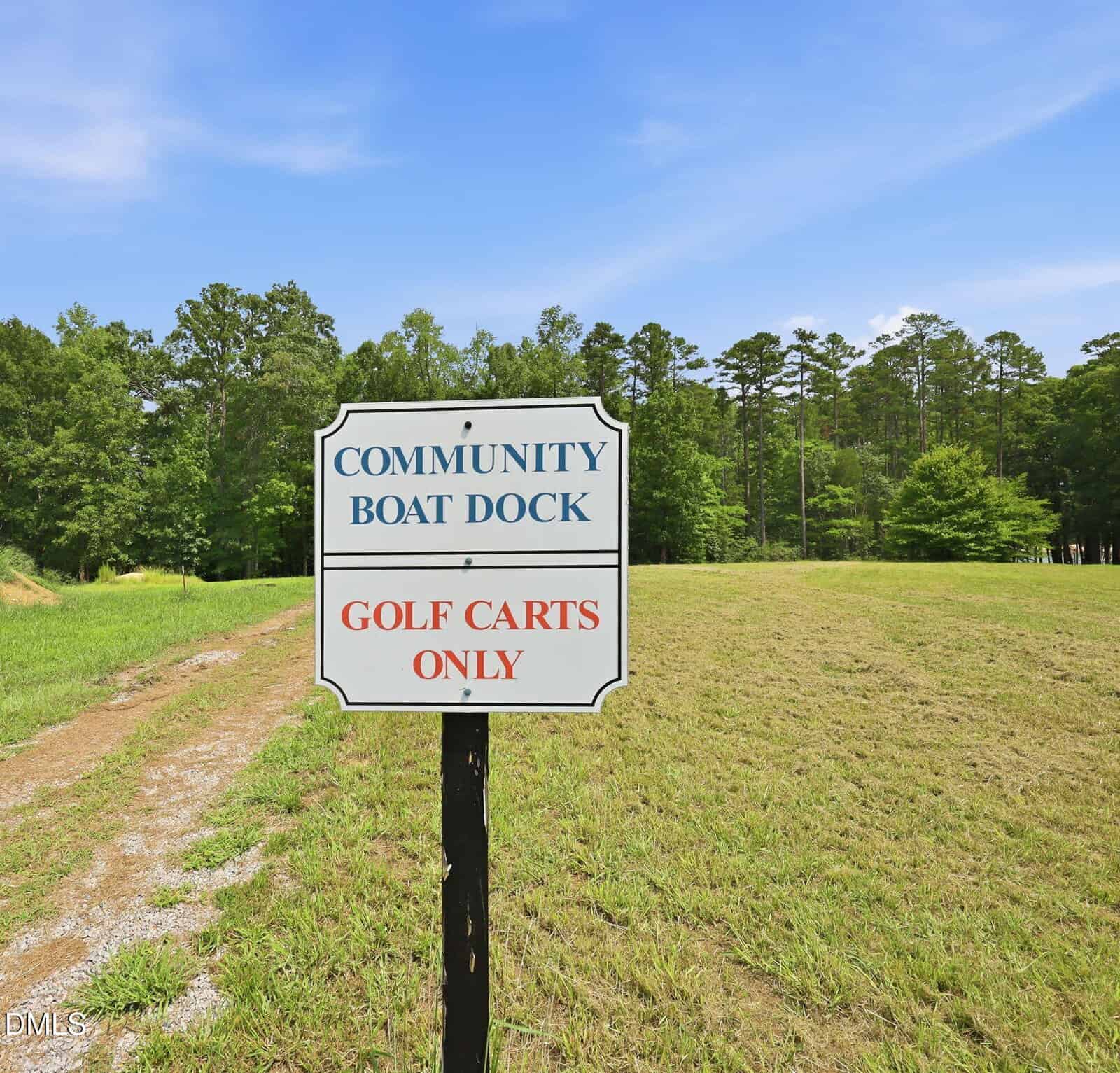 A sign reads “Community Boat Dock” in blue text and “Golf Carts Only” in red text, standing on a grassy area near a gravel path, with trees and a blue sky in the background.