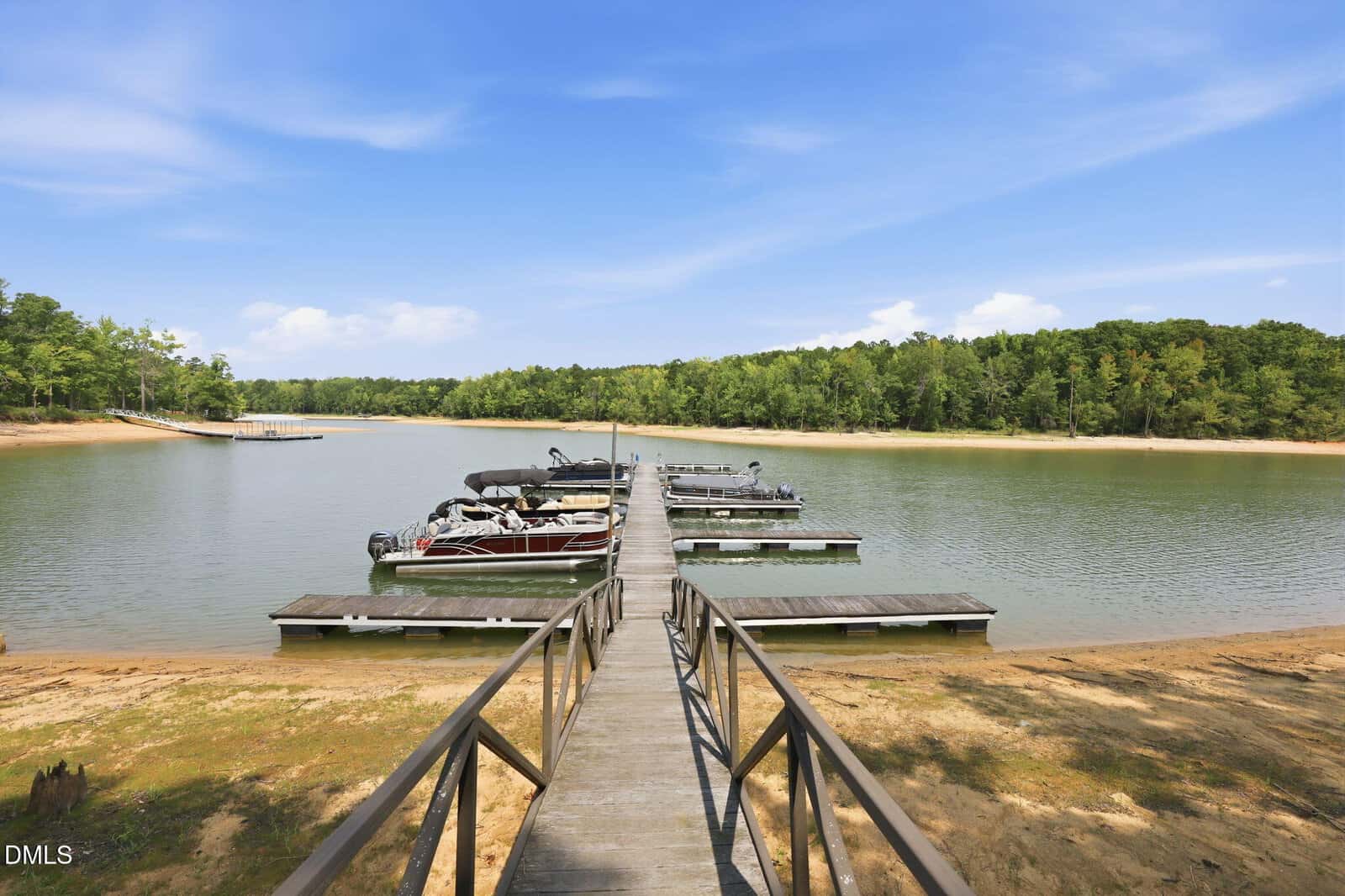 A wooden dock extends over a calm lake with several pontoon boats moored, offering a tranquil setting at 162 Stonewood Loop Lane, Henderson. The shoreline features sandy areas and is surrounded by trees under a mostly clear blue sky.