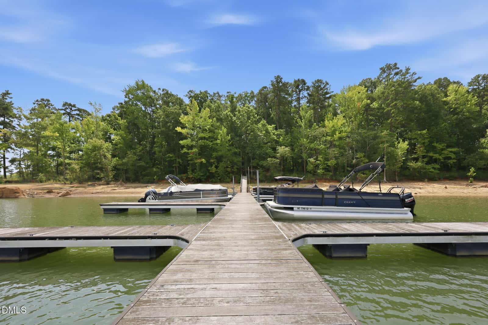 A wooden dock extends into a green lake, with two pontoon boats moored on either side. The shoreline and dense green trees are visible in the background under a blue sky.