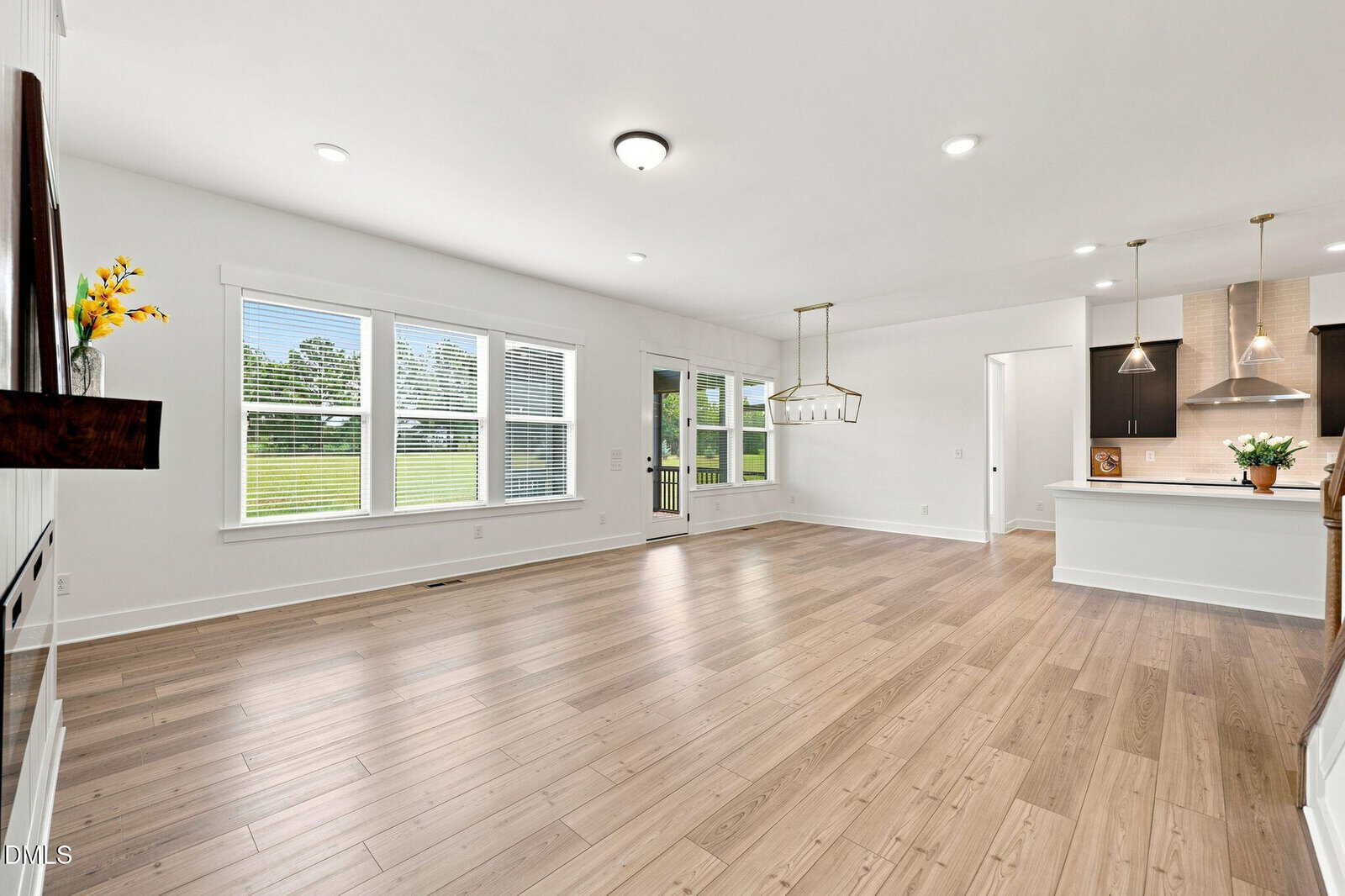 Bright, open living space with light wood floors, large windows, modern lighting fixtures, and a view into a white kitchen with an island and hanging pendant lights. Door leads outside to a grassy yard.