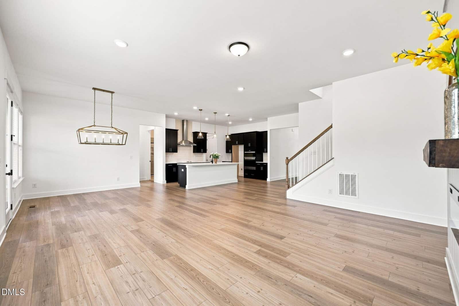 Open-plan living area with light wood floors, white walls, modern kitchen with dark cabinets and island, hanging light fixture, staircase, and yellow flowers in a vase on the right.