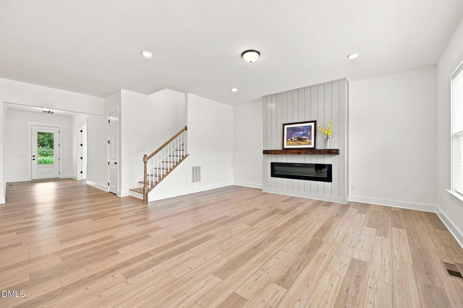 Bright, modern living room with light wood floors, white walls, a staircase, large windows, and a built-in fireplace under a shelf displaying framed art and a vase with yellow flowers.