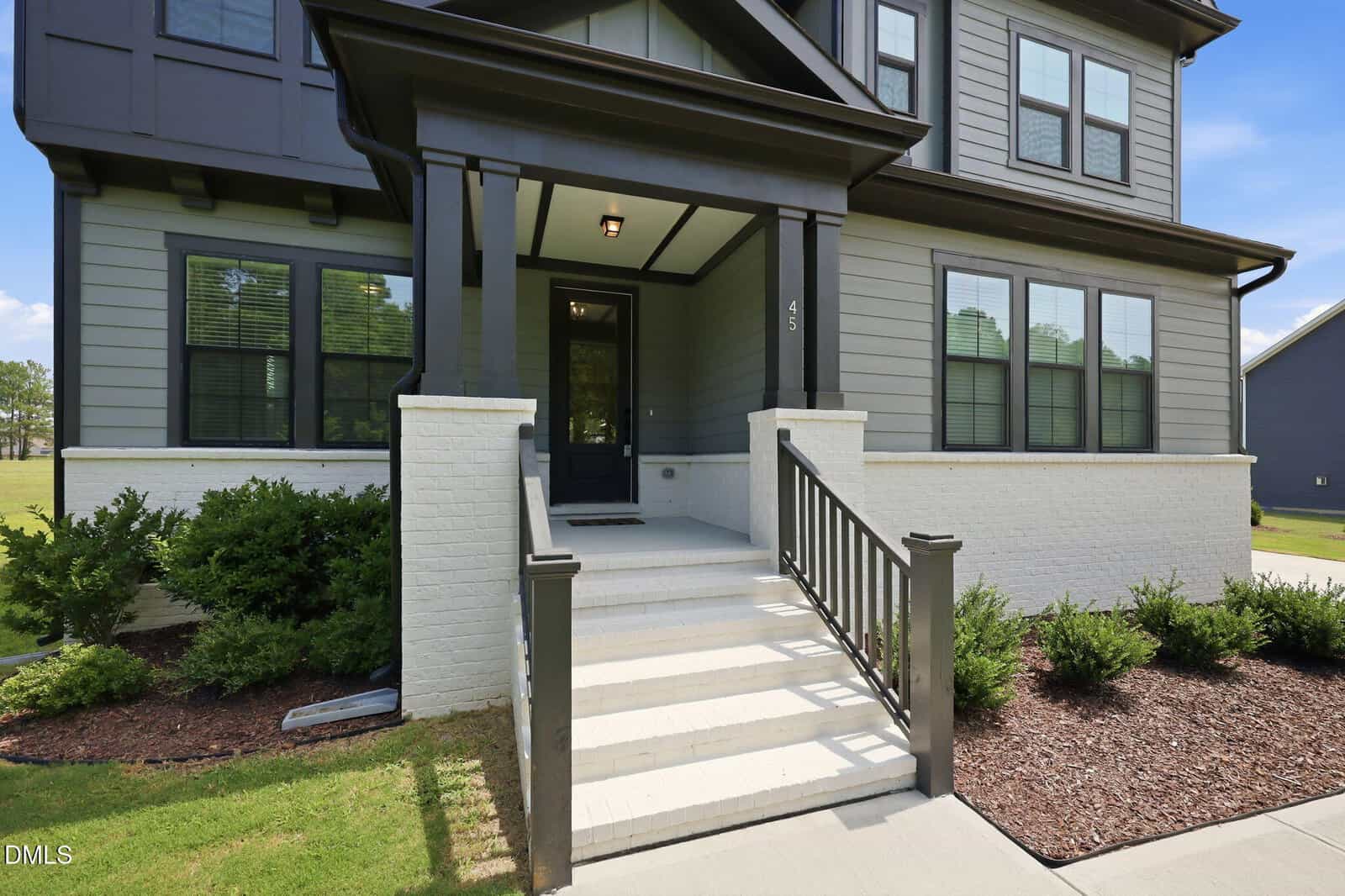 A modern two-story house with gray siding, white brick accents, black trim, a covered front porch, and a set of white steps leading to the front door. Shrubs and mulch line the walkway and the porch.