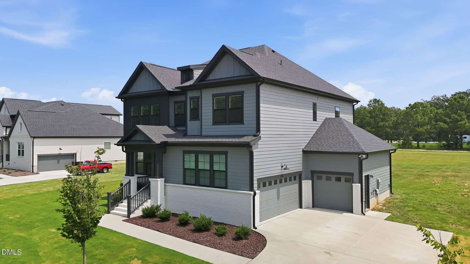 Two-story modern house with gray siding, black trim, and a dark-shingled roof. It has a double garage, driveway, small front steps, and a landscaped yard with grass and a young tree. Other homes and trees are in the background.