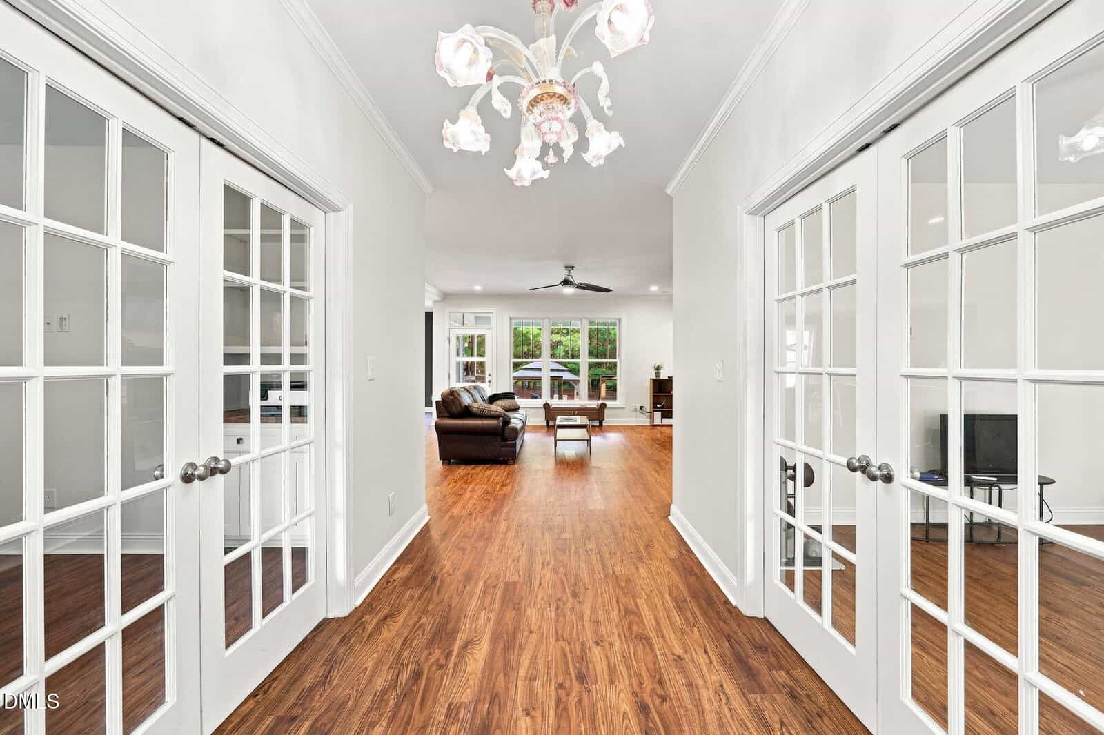 A hallway with double French doors on both sides, wood flooring, and a white chandelier leads to a living area with a brown sofa and large windows.