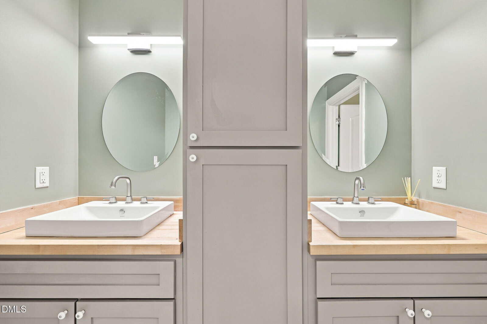 A bathroom with two sinks, each with a circular mirror above, separated by a tall central cabinet. Both sinks have silver faucets and wooden countertops under bright wall lights. Light green walls are visible.