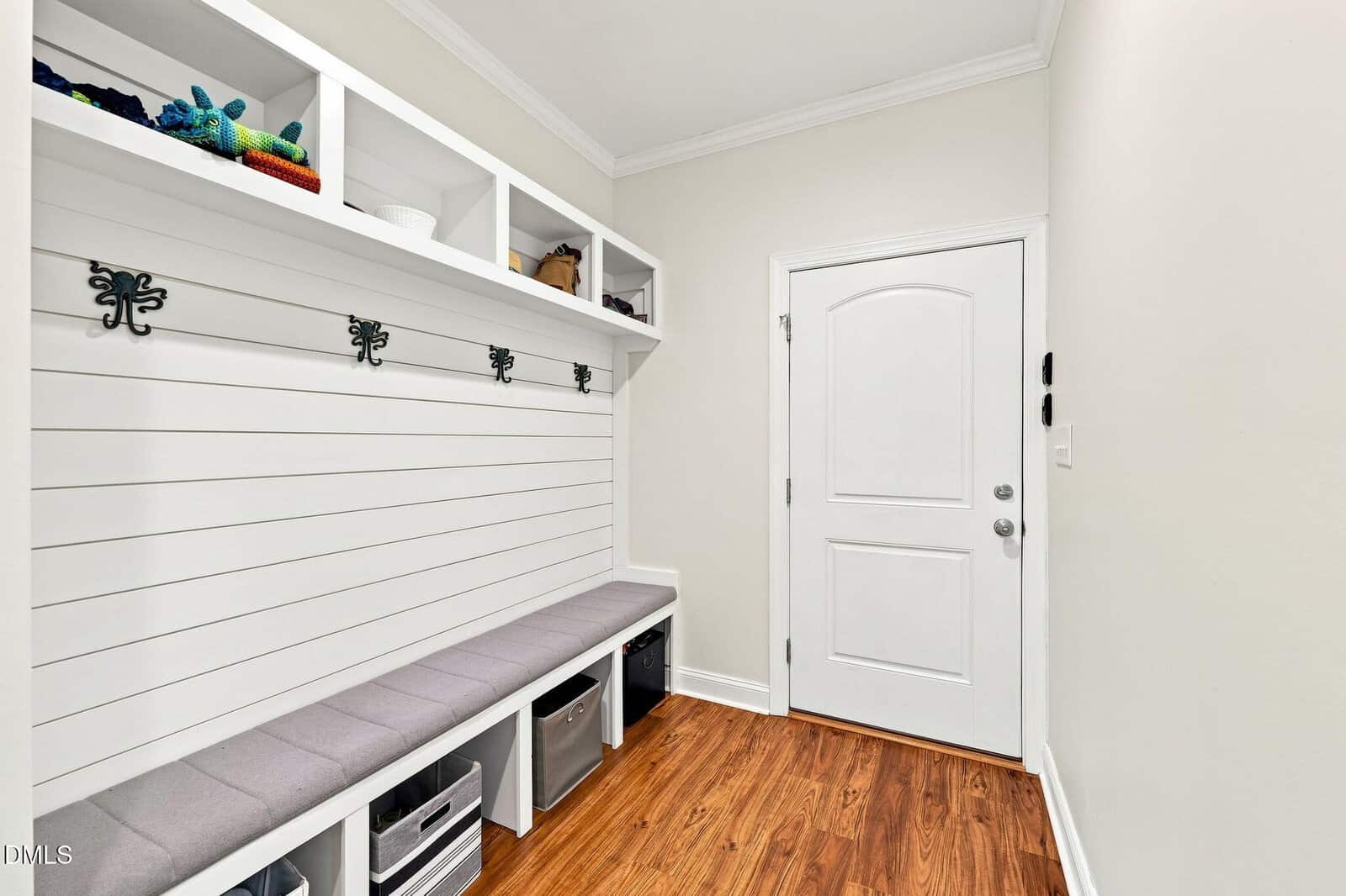 A mudroom with white shiplap walls, bench seating with gray cushions, cubbies below, wall hooks, upper shelves holding baskets, and a white door on the right. The floor is a medium-toned wood.
