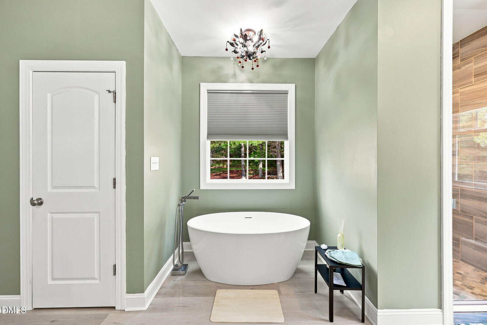 A modern bathroom with green walls, a white freestanding bathtub, a small black side table, a window with a gray blind, a white door, and a contemporary ceiling light fixture.