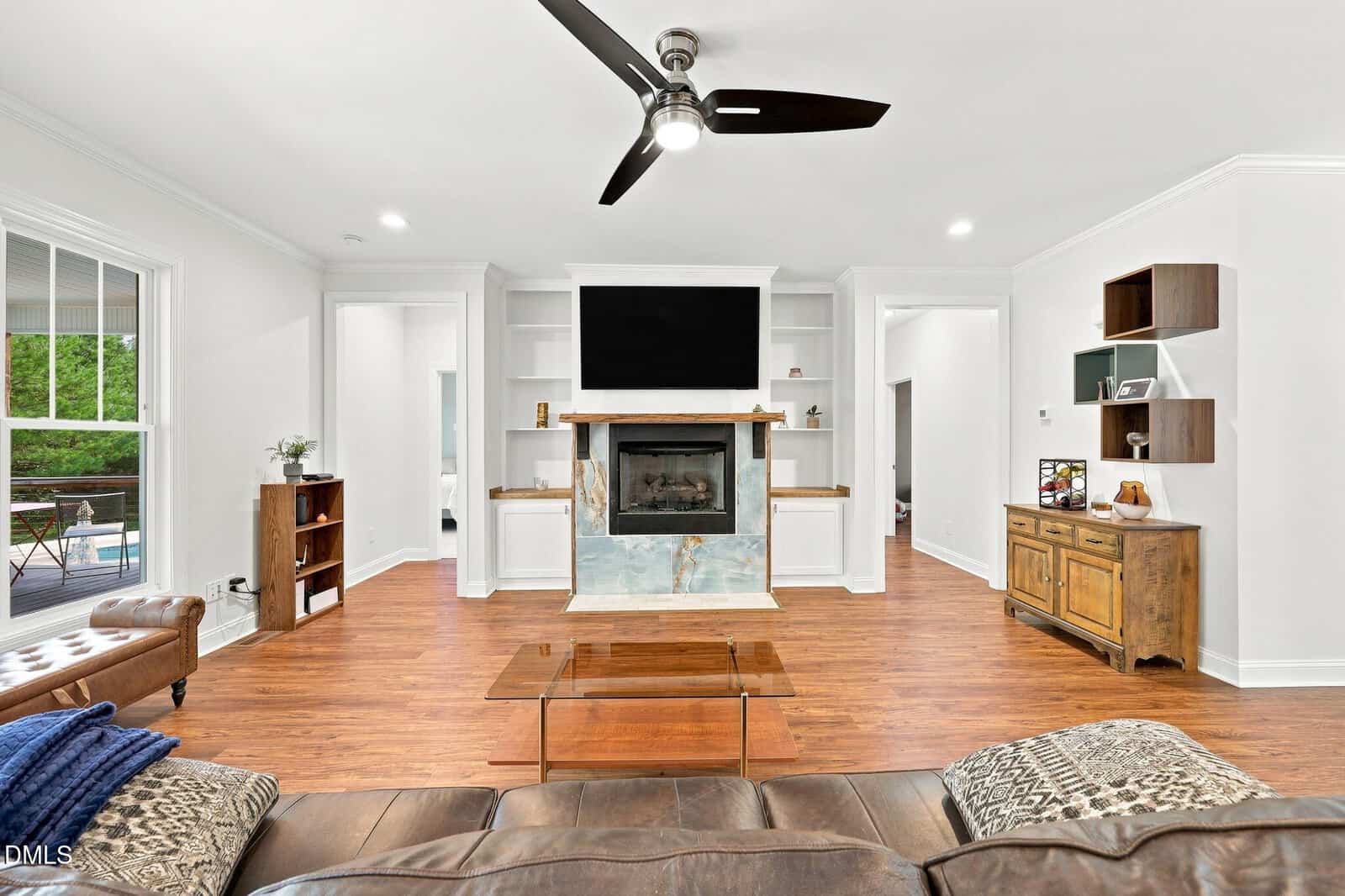 Modern living room with a ceiling fan, fireplace, wall-mounted TV, wooden floors, open shelves, a brown leather sofa, glass coffee table, and a wooden cabinet. Bright, airy space with white walls and natural light.
