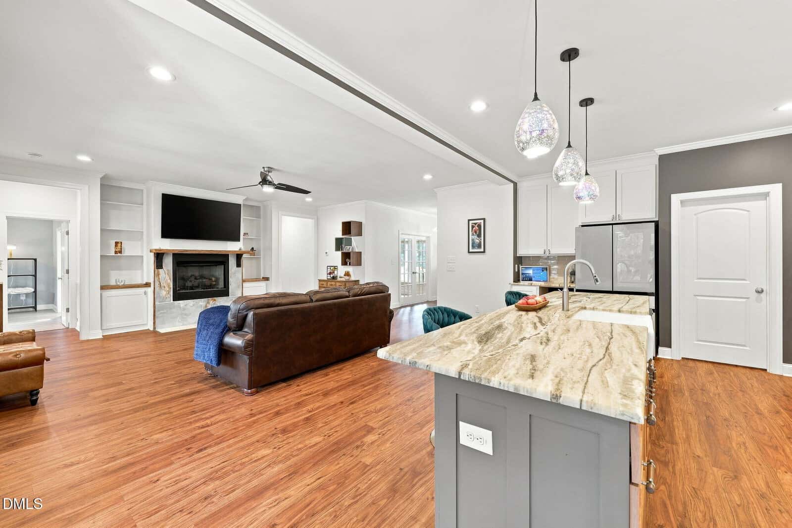 Open living room and kitchen with wood floors, marble island, pendant lights, brown leather sofas, fireplace, built-in shelves, TV above fireplace, and white walls with dark gray accents.