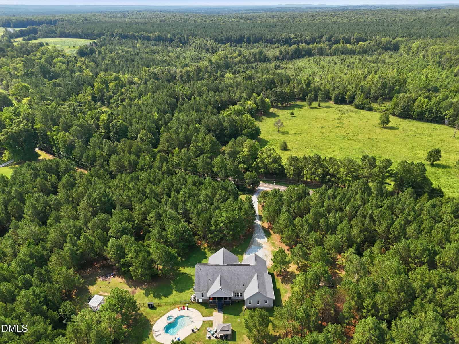 Aerial view of a large house with a backyard pool, surrounded by dense green forest and open fields under a clear sky.