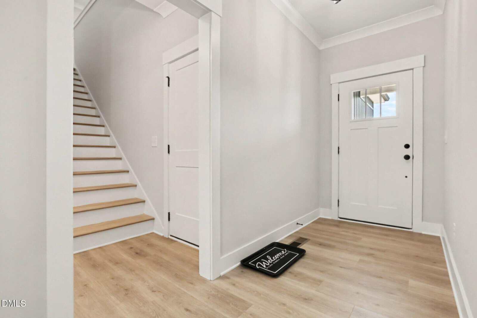 A modern entryway with light wood floors, white walls, a white front door with glass panes, stairs on the left, and a doormat that reads “Welcome.”.